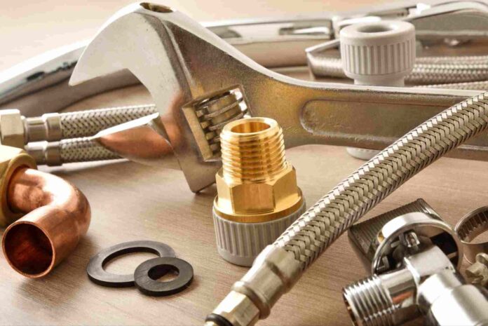 An overhead shot showing a variety of plumbing supplies, including a large adjustable wrench, brass and chrome fittings, a flexible metal hose, and rubber washers, arranged on a light-colored wood backgroun